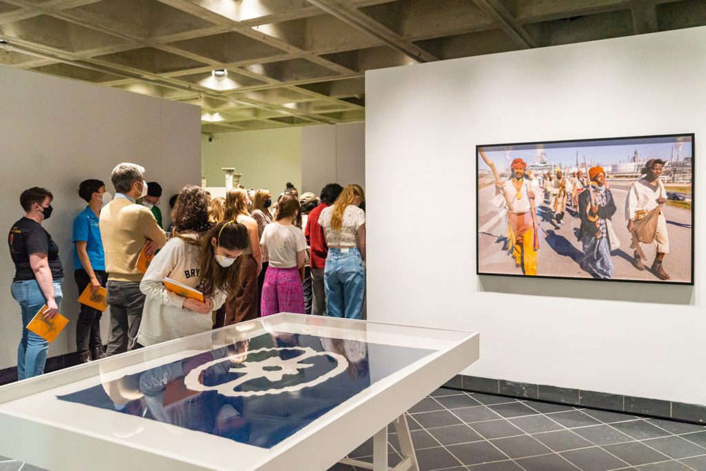 A crowd of students walk through the gallery with the SRR flag and photo from the Slave Rebellion Reenactment visible.