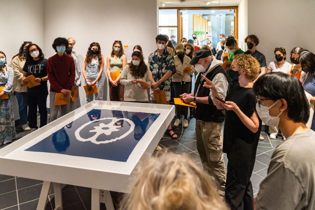 A crowd, including John Muse and curator Sally Berger, stand around a large table containing a blue and white SRR flag