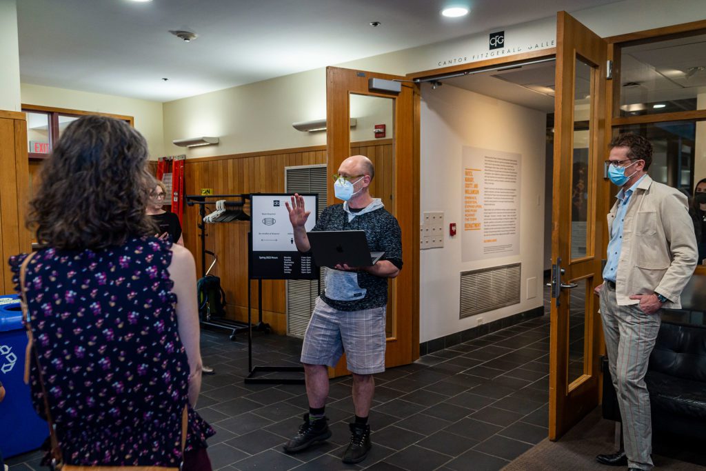 John Muse wearas a mask and holds a laptop standing at the entrance to the gallery.