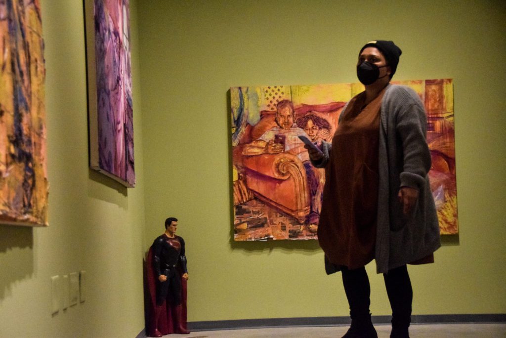 A masked visitor to the gallery stands in the corner of the gallery near the Superman figurine and looks at three different paintings.