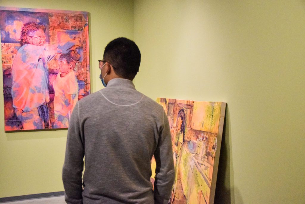A gallery visitor looks at a painting of a woman cooking in a kitchen and another of a parent measuring a child against the wall.