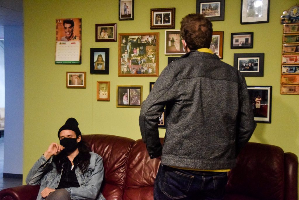 One visitor sits on the red couch in the gallery while another stands and looks at a wall of family photographs