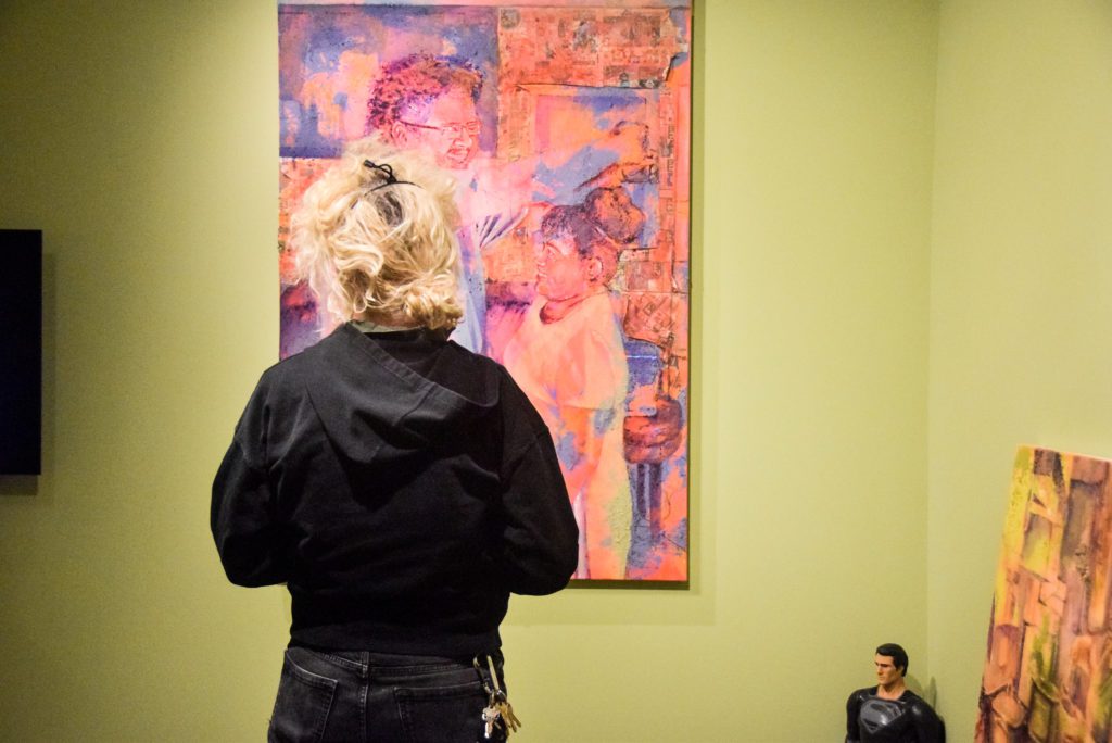 A visitor stands in front of a pink and purple painting of a parent measuring a child against the wall.