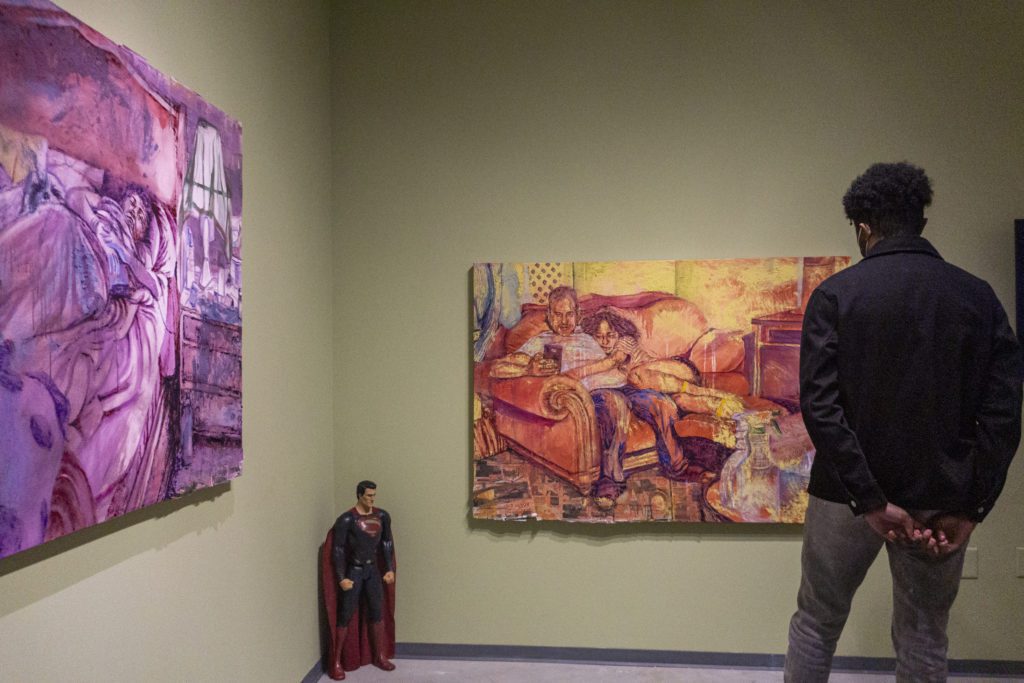 An attendee looks an orange and red painting of two people sitting on a couch while one looks at a cell phone. In the corner of the gallery is a Superman figurine.