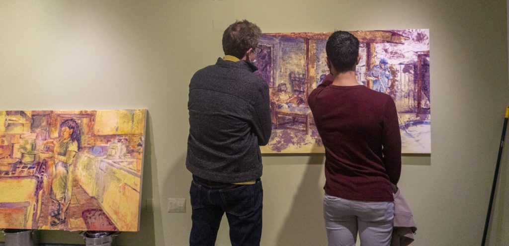 Two attendees stand in front of two of Morales' pantings of home, including one propped up on pots and pans