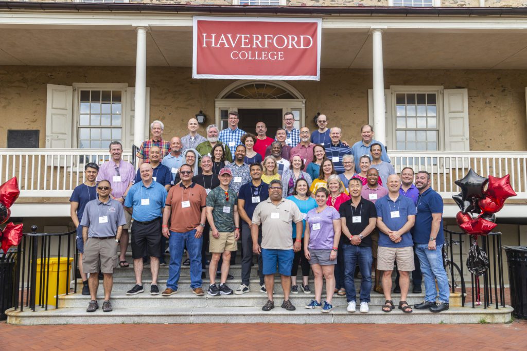 Roughly 40 people in shorts and t-shirts pose on the Founders steps under a Haverford College banner.