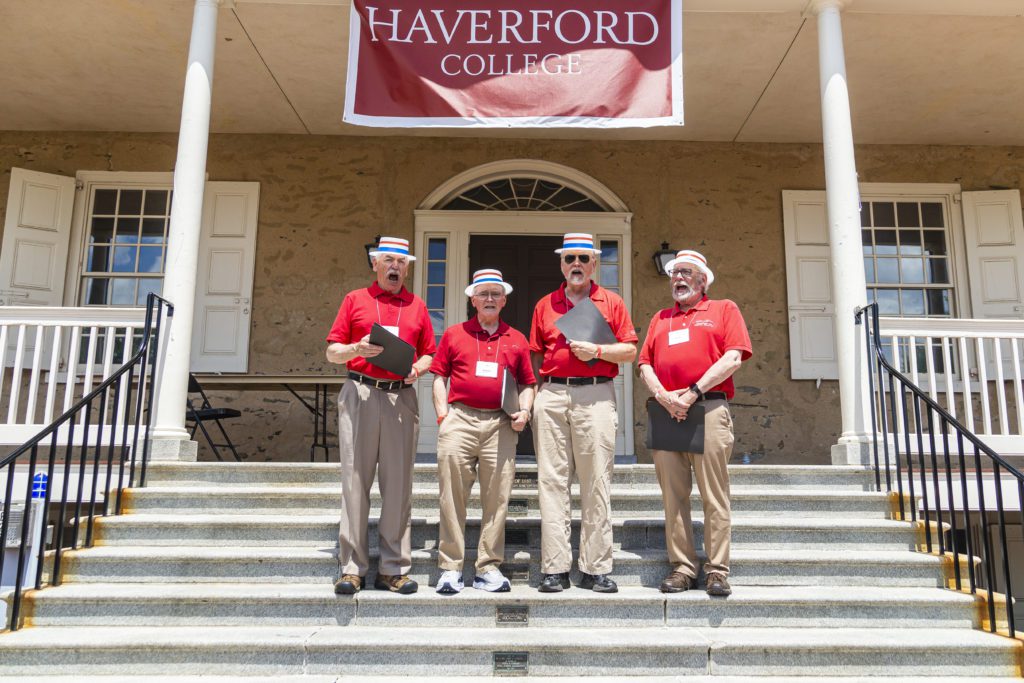 Four singers in red shirts and boater hats sing under a Haverford College banner on Founders steps.