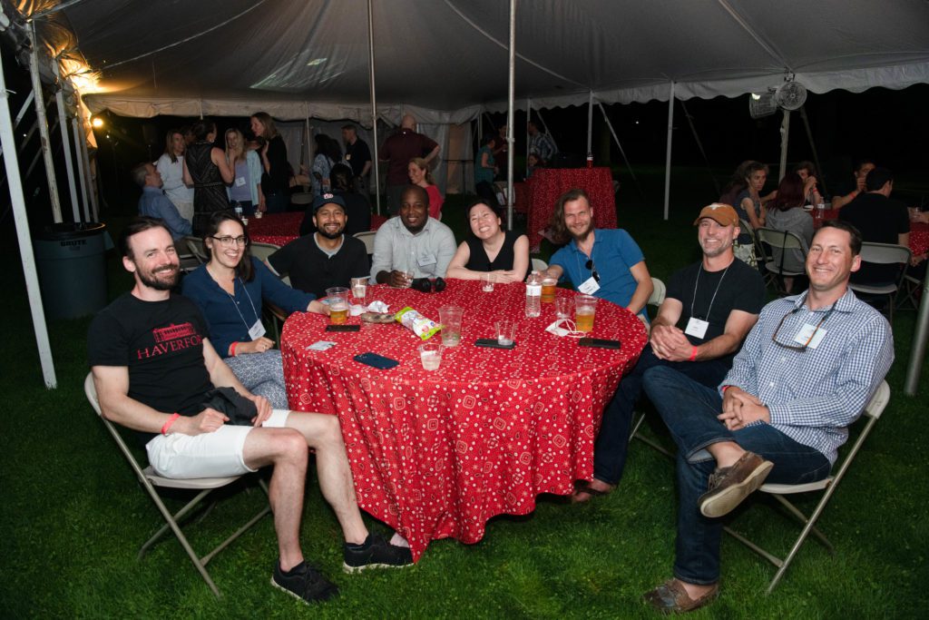 Eight classmates smile, seated around a round table that is full of drinks and phones