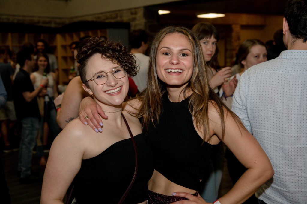 Two young alumni, both dressed in black, smile with their arms around each other.