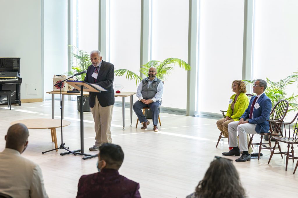Harold Weaver '56 P'03 stands at the microphone with three people sitting behind him and light streaming in through the windows at the back.