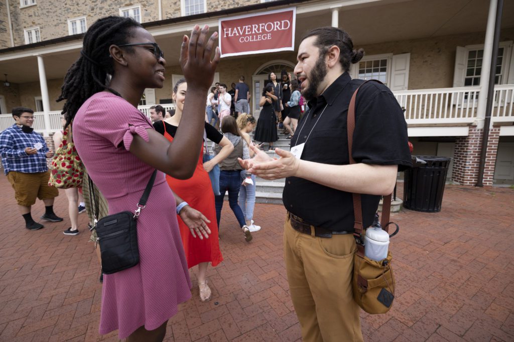 Two alumni talk animatedly to one another in front of a crowd of alumni on Founders porch under a Haverford College banner.