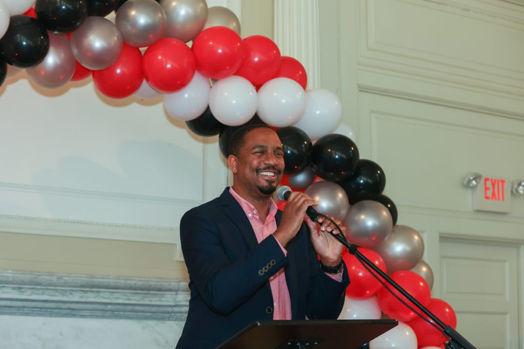 John McKnight smiles at the podium as he speaks in front of a balloon arch