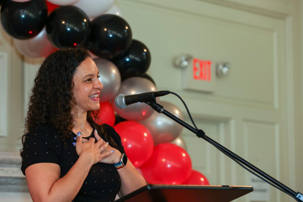 Christina Rose smiles and touches her heart while speaking at the podium.