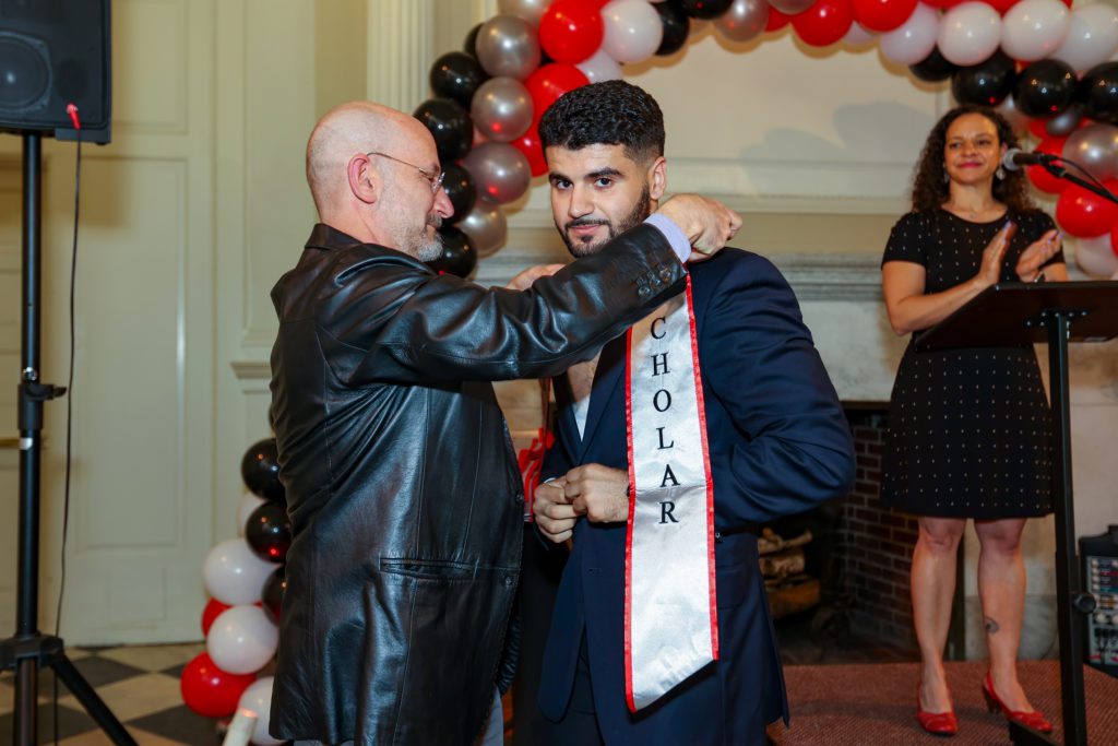 Jeff reaches over Ali's head to put his sash on with Christina Rose clapping in the background