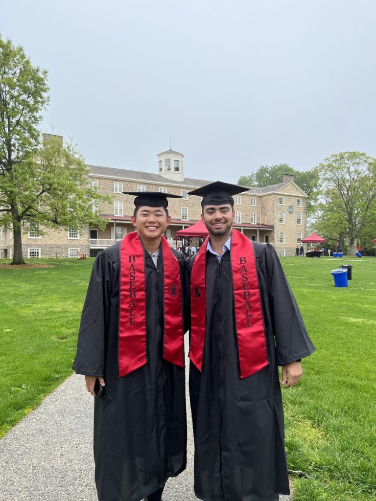 Two students in caps and gowns wear red "baseball" sashes in front of Founders before Commencement.
