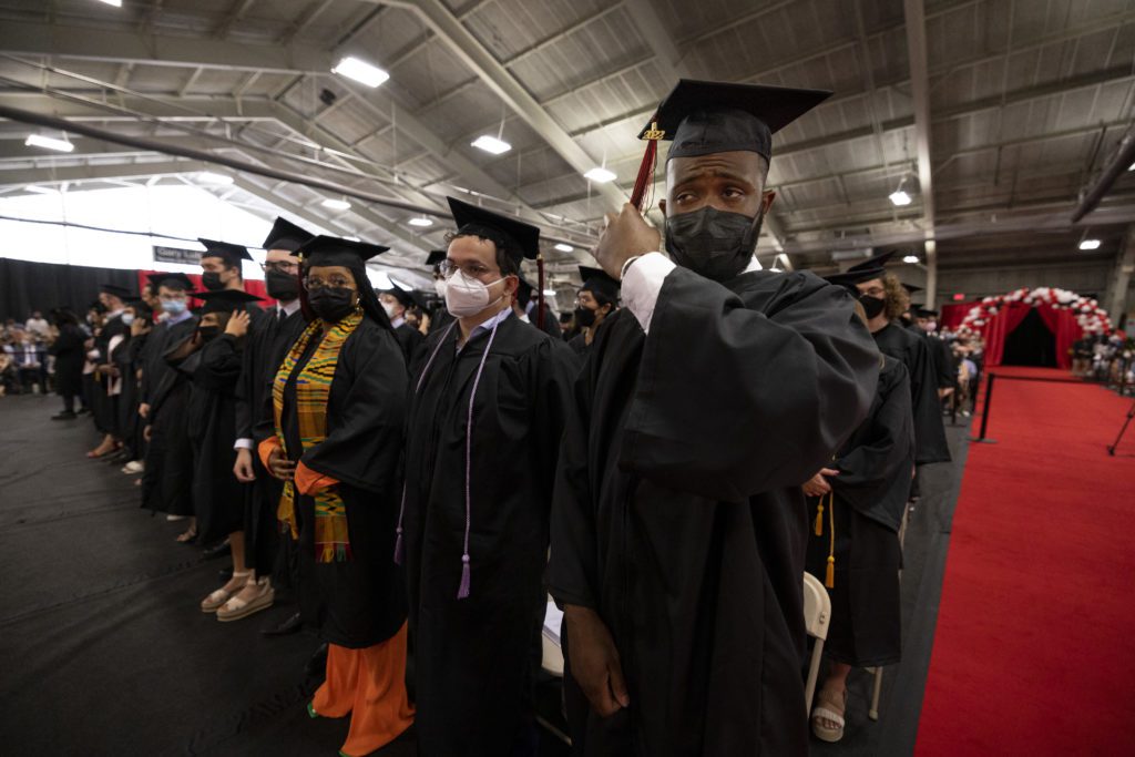 A group of masked students in caps and gowns stand and prepare to move their tassels.