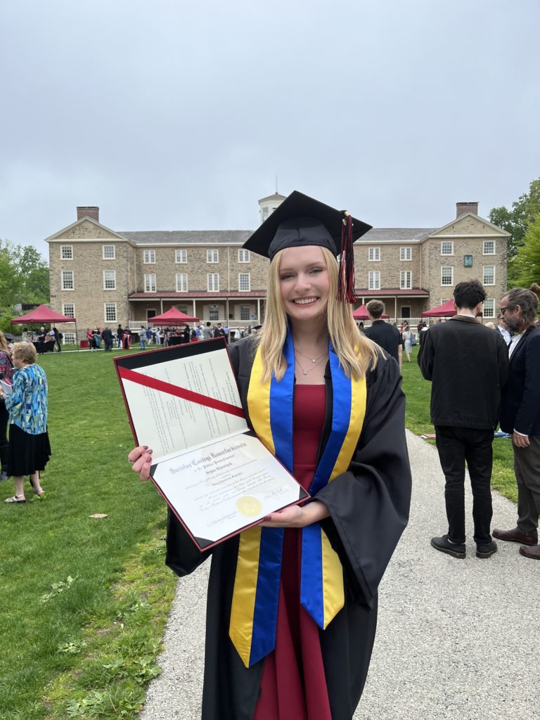Iryna in a cap and gown and blue and yellow sash holds up her diploma in front of Founders Hall