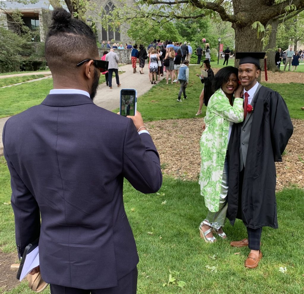 A student in a cap and gown poses with a family member for someone taking their photo with a phone.