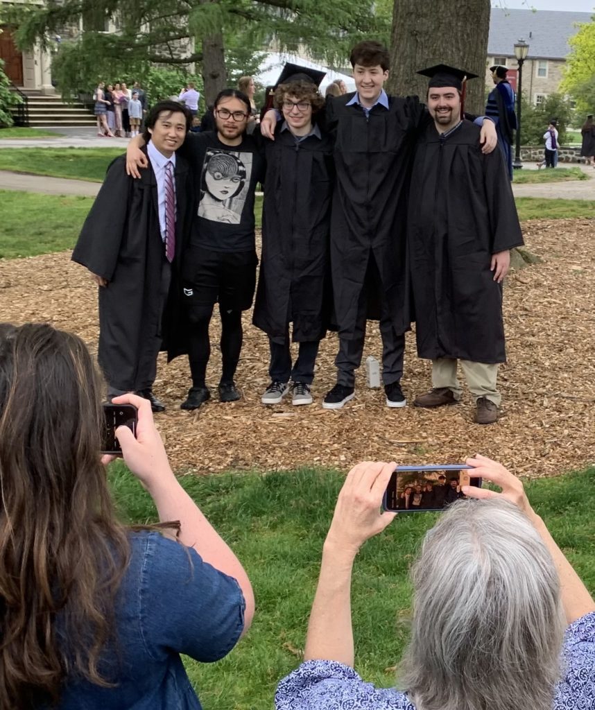 Five people in caps and gowns pose with arms around each other as family members take cell phone photos
