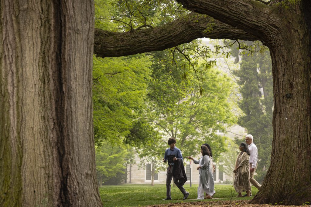 A student holding a cap and gown is trailed by his family as they walk through the trees.