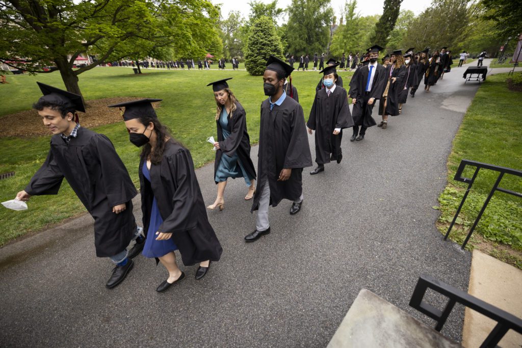 Students in caps, gowns, and masks walk two-by-two from Founders to the Field House