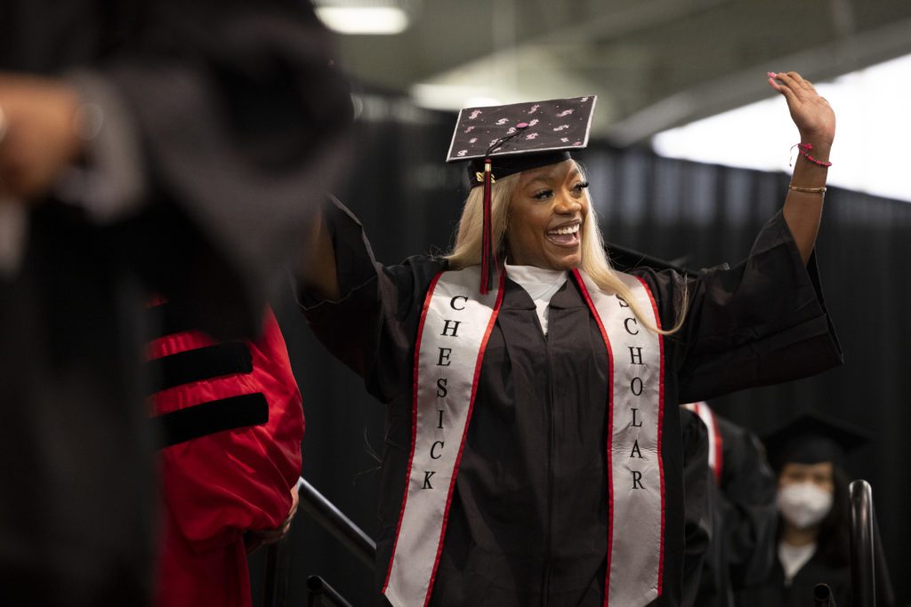 A student in a decorated cap with a Chesick Scholar sash waves at the audience as she approaches the stage to get her diploma from the president.