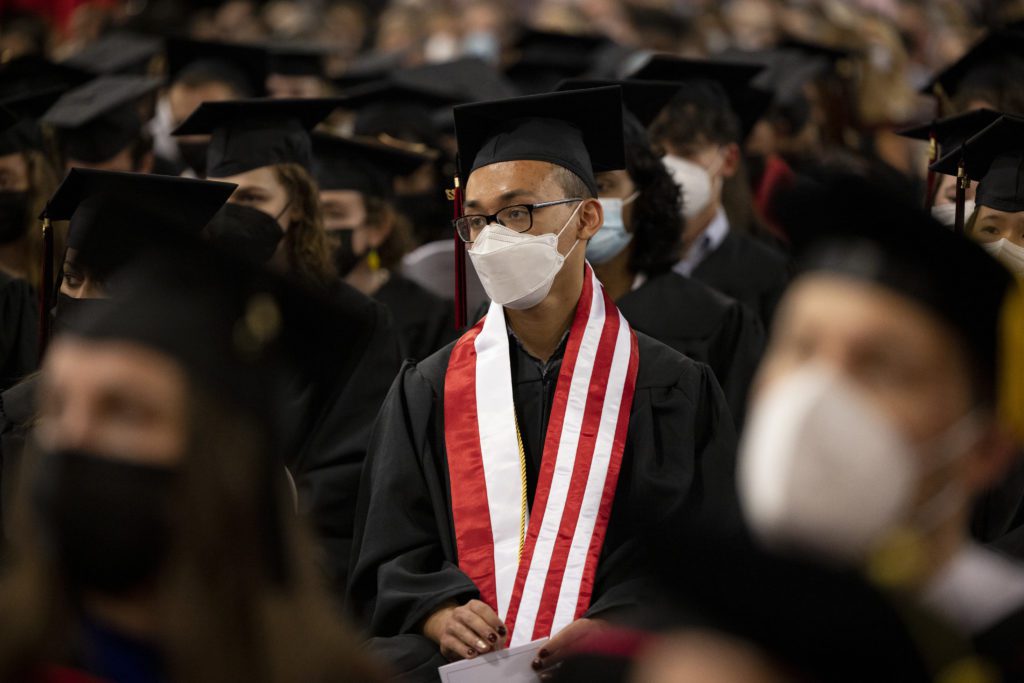 A student in a mask and a red and white striped sash sits among a sea of black robes and caps