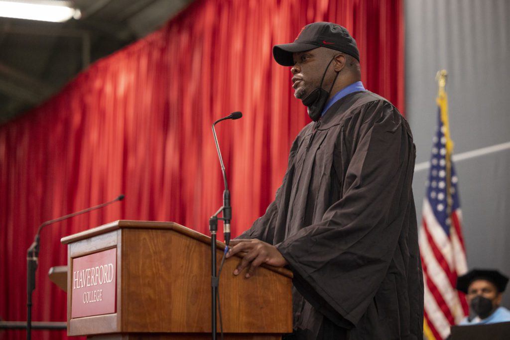 Charles wears a cap with his mask pulled down and speaks at the podium.