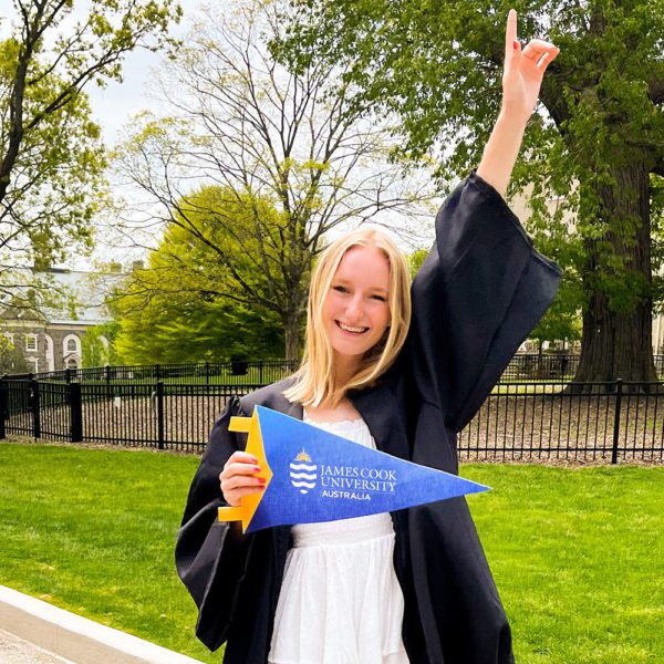 Genevieve Dallmeyer-Drennen '22 stands on Haverford's campus, wearing her graduation gown, holding a James Cook University pennant.