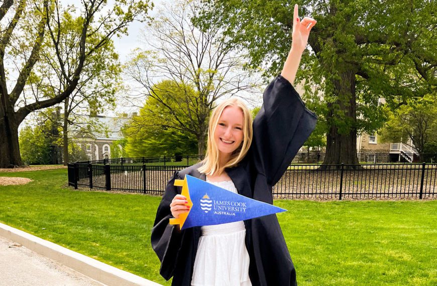 Genevieve Dallmeyer-Drennen '22 stands on Haverford's campus, wearing her graduation gown, holding a James Cook University pennant.