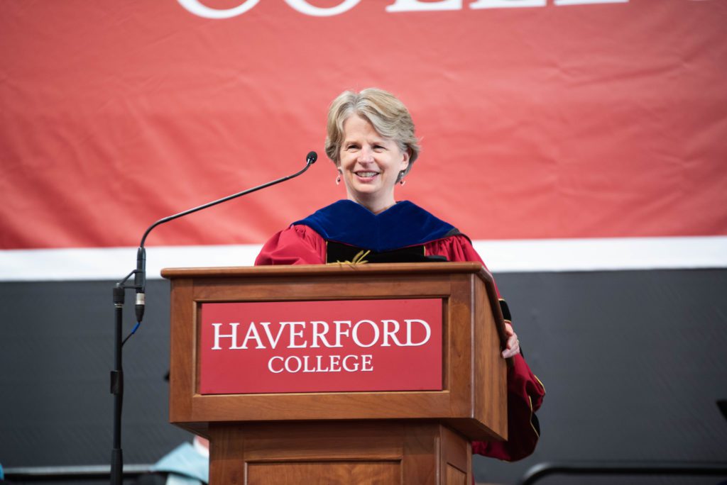 President Raymond wears a red robe and blue hood and speaks at a podium.