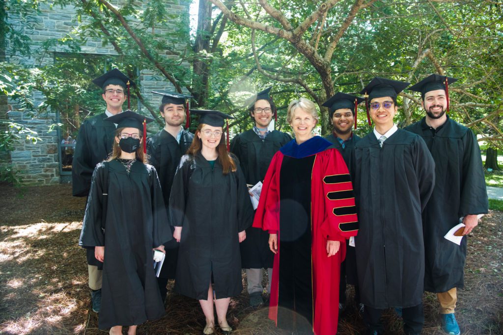 President Raymond and her red robe stands out among a sea of black robes as she poses, smiling, with eight graduates.