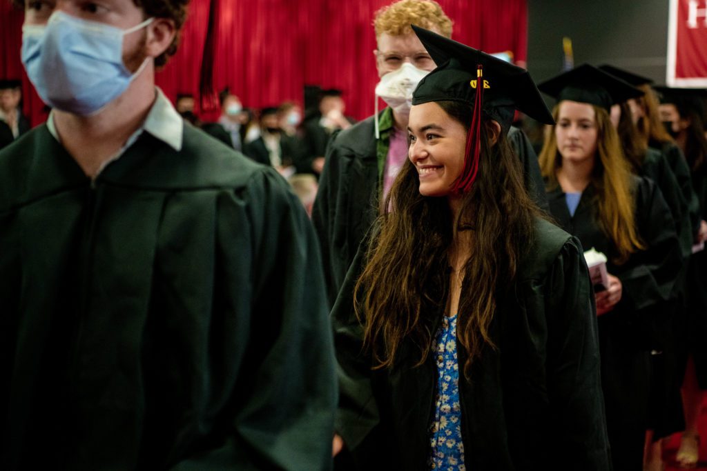 Caroline smiles in her cap and gown and Lev walks behind her in a mask.