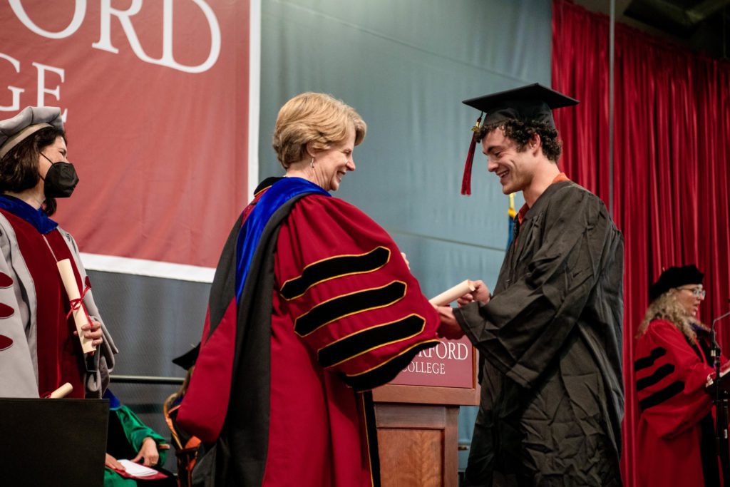 A graduate in a black cap and gown takes a scroll from President Raymond onstage