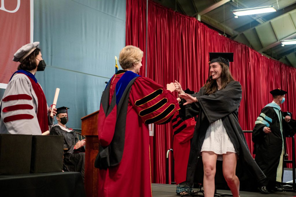 A graduate in a white mini dress under her black gown fist bumps President Raymond in her red robe as she walks across the stage.