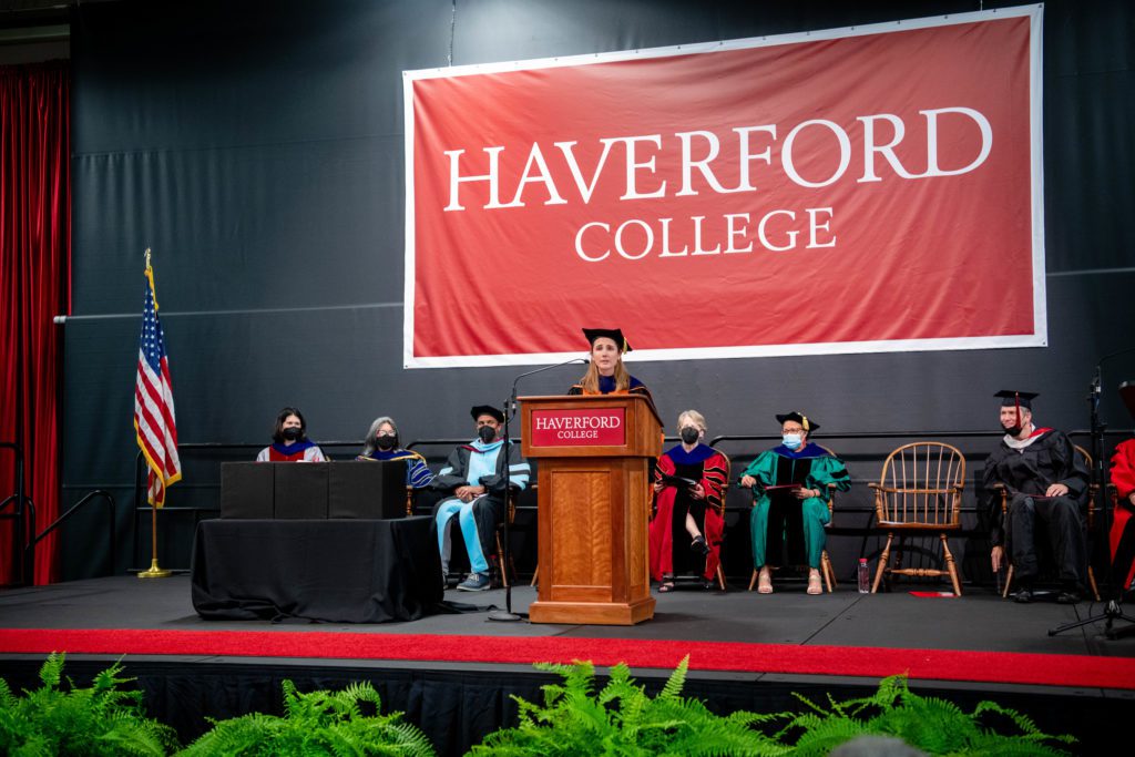 Lindsay wears a black cap with a gold tassel and stands in front of a line of faculty and administrators in their academic regalia under a giant Haverford College banner onstage.