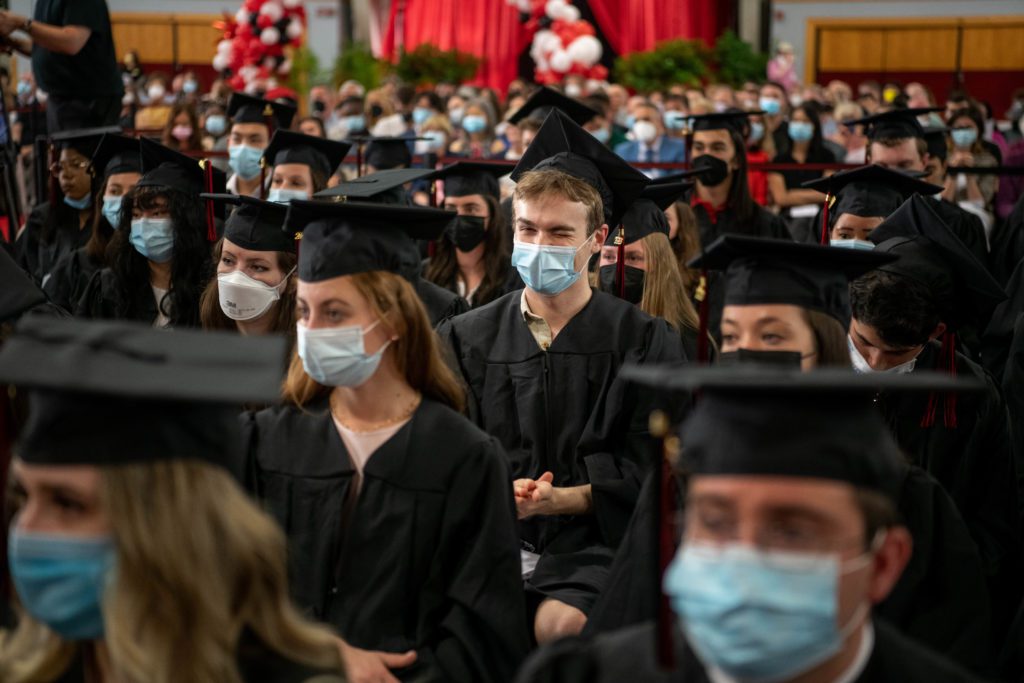 A graduate in a sea of masked graduates in caps and gowns winks at the camera.