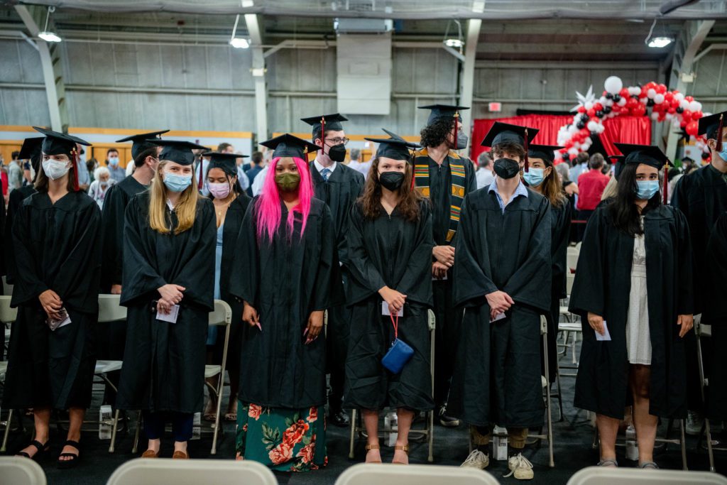 About 10 members of the class stand at their seats in their black caps and gowns.