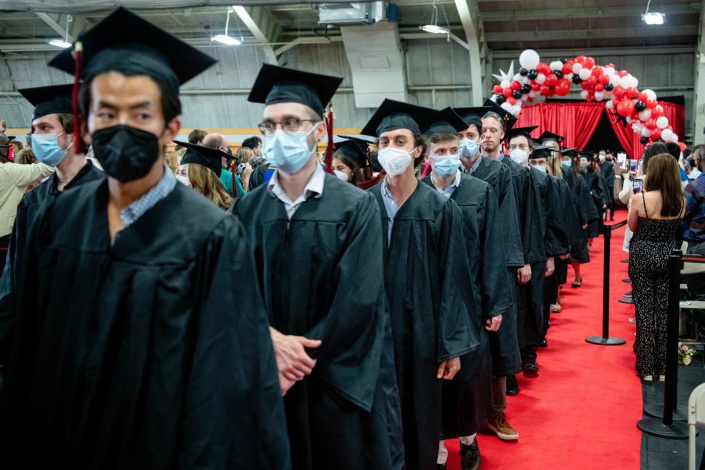 Graduates in their caps and gowns walk in a line down a red carpet through a balloon arch.
