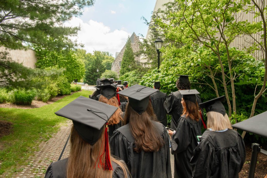 A view from behind the processional of black robed graduates through the trees.