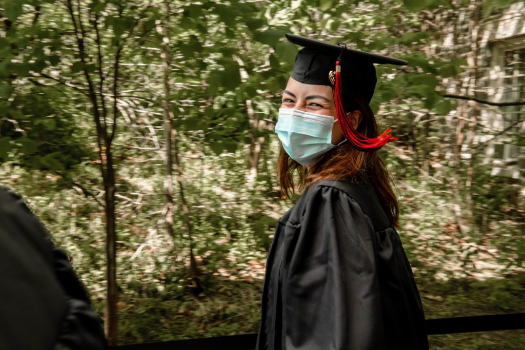 A masked graduate in a cap and gown smiles over her shoulder.