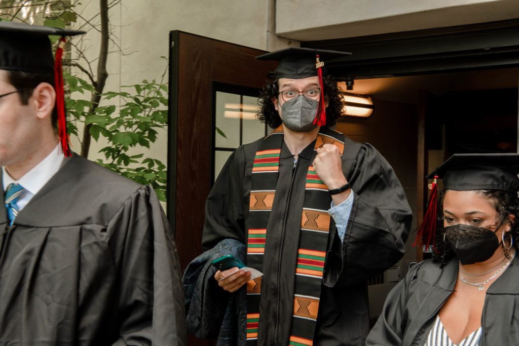 Joe wears his cap and gown and stole and fist pumps at the camera.