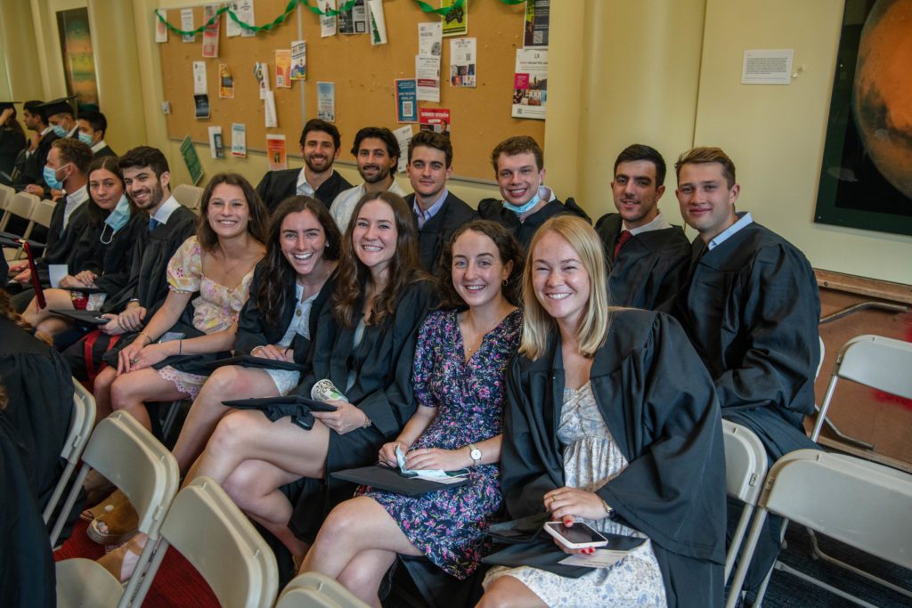 14 members of the Class of 2020 sit in rows in their black graduation gowns waiting to line up for the processional.