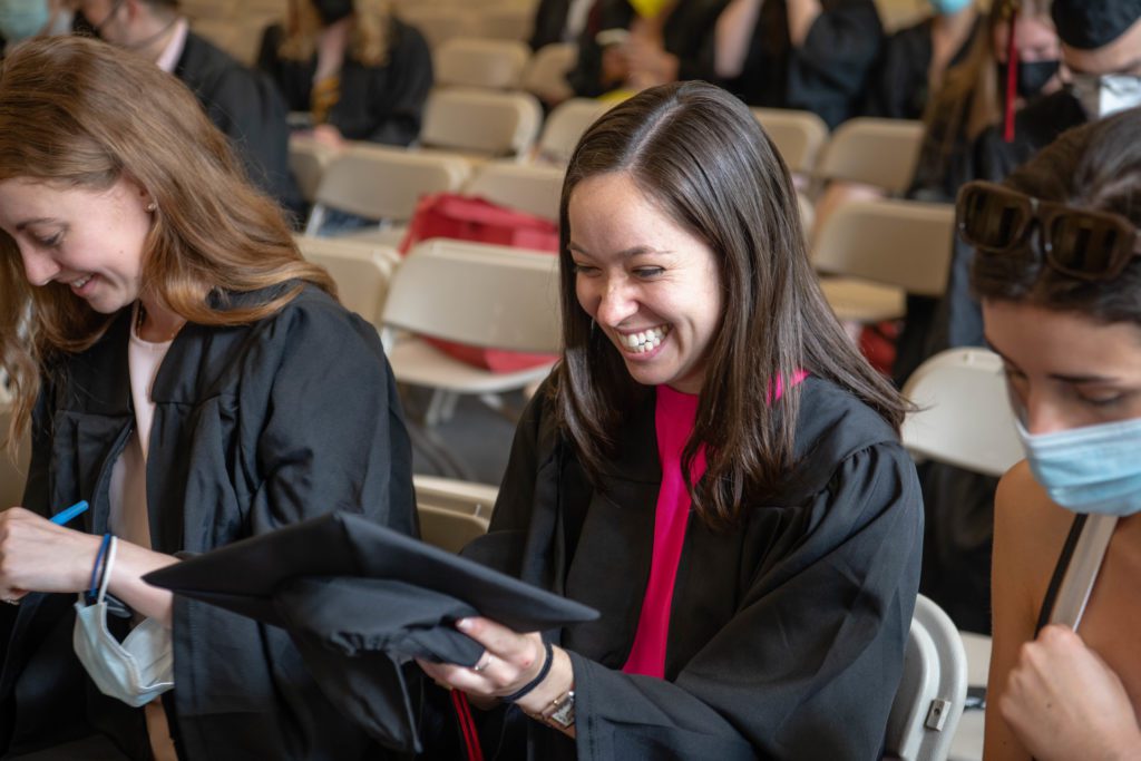 Three graduates sit in the staging area in their black gowns and one laughs at her cap