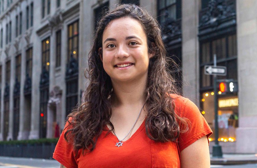 Anne wears an orange shirt and smiles on a Manhattan street