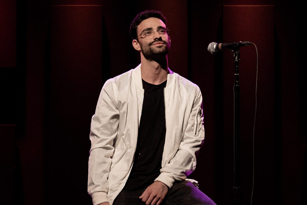 Yuval leans on a high stool onstage with a velvet curtain behind him