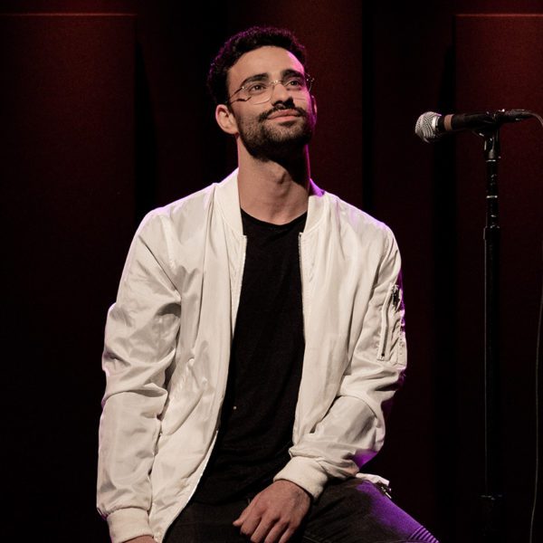 Yuval leans on a high stool onstage with a velvet curtain behind him