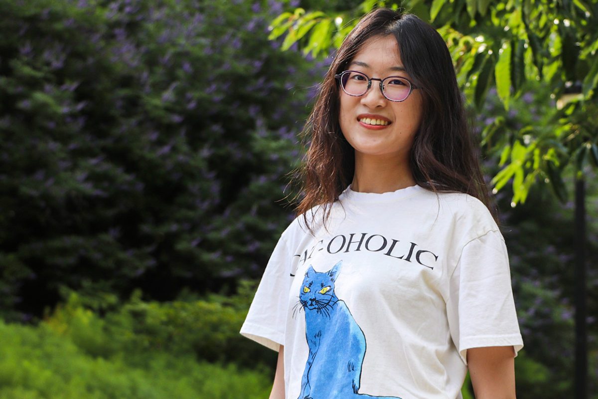 Jianing Mu '22 stands in front of a green background of trees and plants, smiling. Photo by Aby Isakov '24.