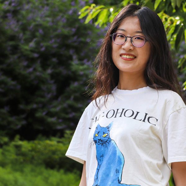 Jianing Mu '22 stands in front of a green background of trees and plants, smiling. Photo by Aby Isakov '24.