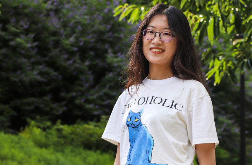 Jianing Mu '22 stands in front of a green background of trees and plants, smiling. Photo by Aby Isakov '24.